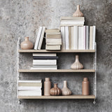 String Pocket Regal Eiche/Weiß mit Büchern und Vasen an einer Betonwand